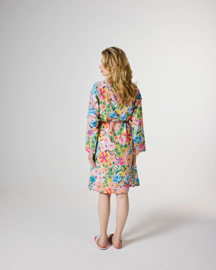 A woman stands with her back to the camera wearing a colorful floral robe with a matching tie belt, set against a plain light gray studio background.