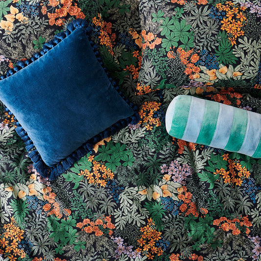 Decorative pillows on a floral and greenery patterned fabric background. Fabric is black base with lots of different green leaves and some blue and orange flowers.