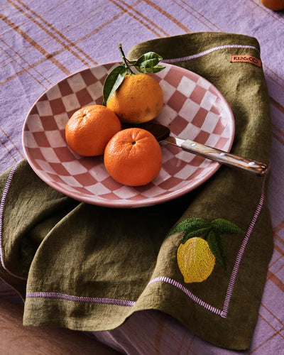 Embroidered khaki green napkin with yellow lemon embroidery, laid underneath pink gingham plate and mandarins.