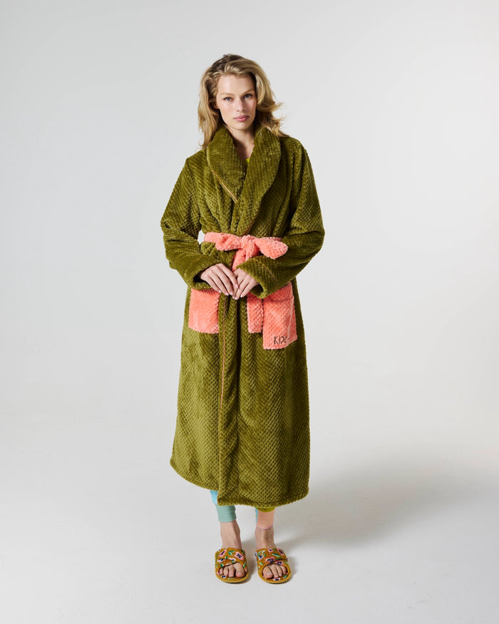 A woman modeling a plush olive green robe with a coral pink tie belt and matching pocket details, standing against a light gray studio background.