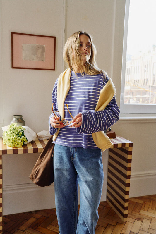 A woman stands indoors wearing a blue and white striped long-sleeve top, blue jeans, and a beige cardigan draped over her shoulders, holding a brown tote bag.