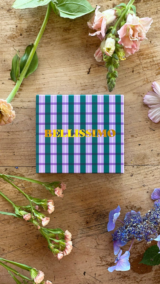 Bellissimo written across front of box in gold leaf. boxed plaid pattern in green and lilac. timber table and various flowers sat around.