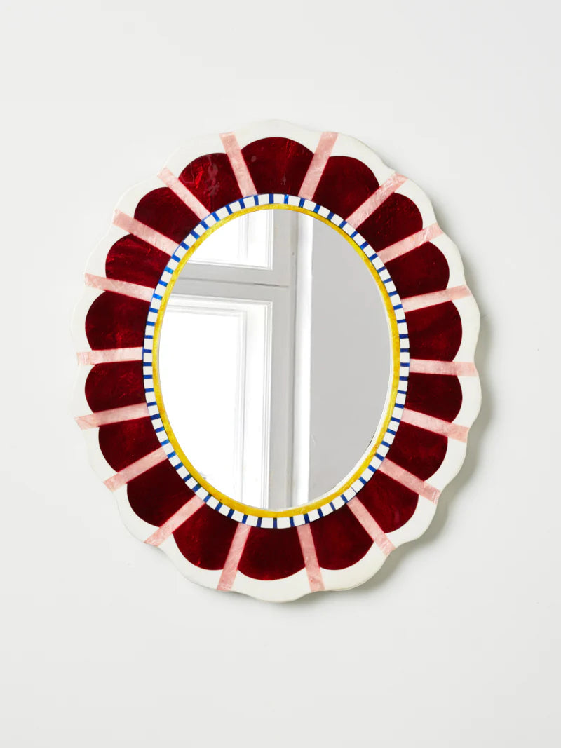 Decorative oval mirror with a colorful frame on a white wall. Colours include wine red, pink, mustard and blue details.