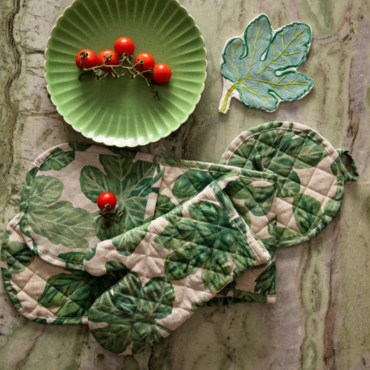 Product shop of leaf-patterned potholder, oven mitt and with a green plate and tomatoes on a stone surface.