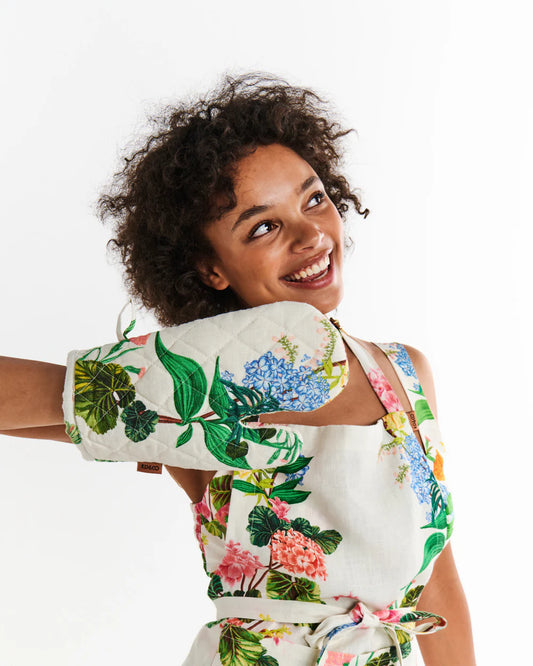Woman wearing a floral apron with a white background and colourful floral pattern with hydrangea flowers and greenery, woman also wears and holds hand out to show oven mitt in matching print.