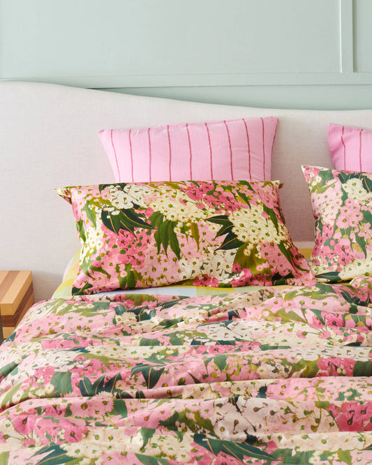 Bed filled with bush blossom quilt cover and matching pillowcases, with pink and red stripe euro pillowcases sat at back of bed. Natural bedhead and green wall.