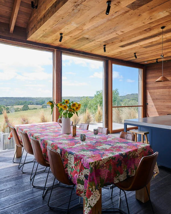Bush blossom floral colourful print laid out on table with leather chairs sat around, in background beautiful view of hills and trees. Timber lined walls and roof.