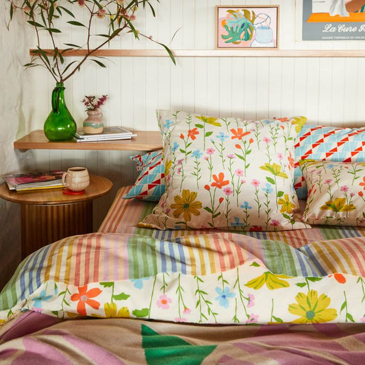 Bed with colorful floral and striped bedding, side table with green vase and books, wooden shelf with plants and decor. Styled by Sage and Clare brand.