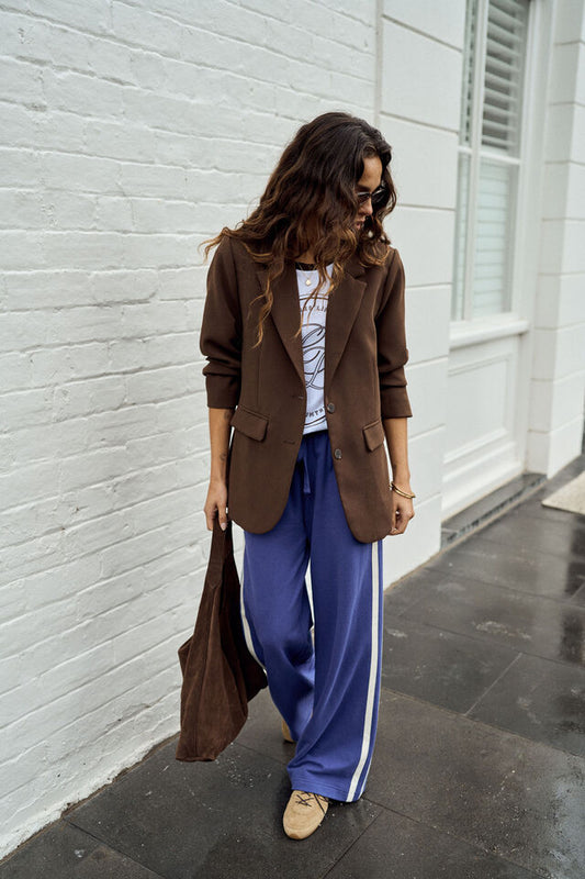 A woman stands on a sidewalk next to a white brick wall wearing a brown blazer, a white graphic t-shirt, wide-leg blue track pants with white side stripes, and tan loafers. She is holding a brown tote bag.