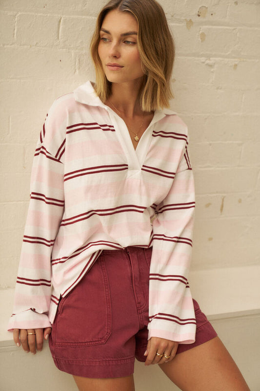 A woman wearing a light pink long-sleeve half-zip pullover with dark red horizontal stripes, paired with burgundy shorts, posing against a white painted brick wall.