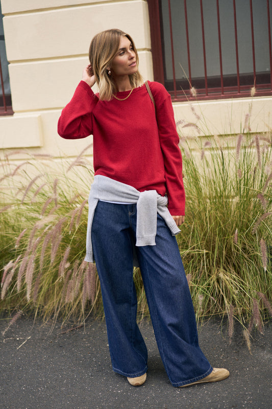 Woman wearing a red sweater and blue jeans standing outdoors with tall grasses in the background.
