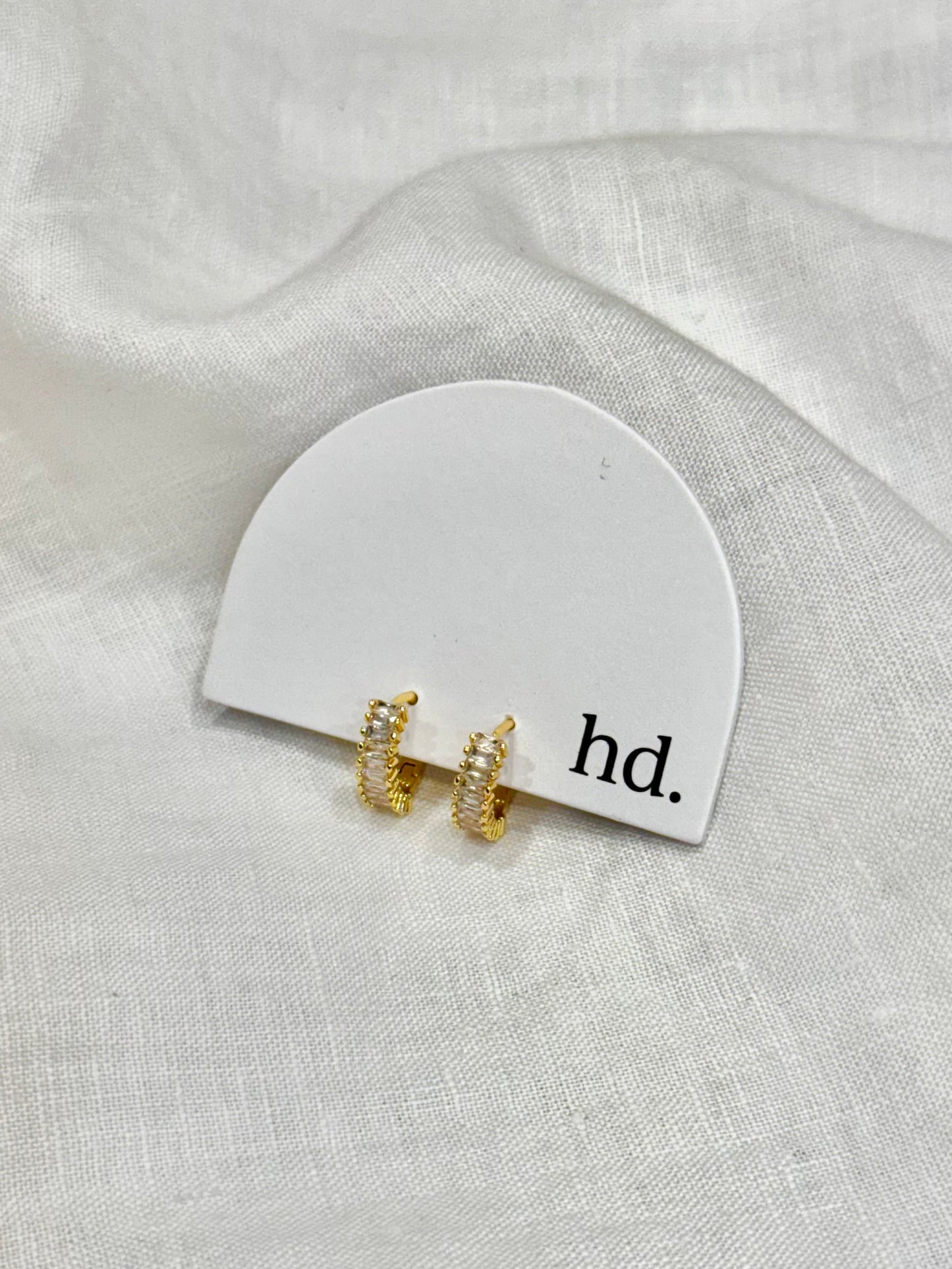 Gold earrings on a white card with 'hd.' text on a light gray fabric background