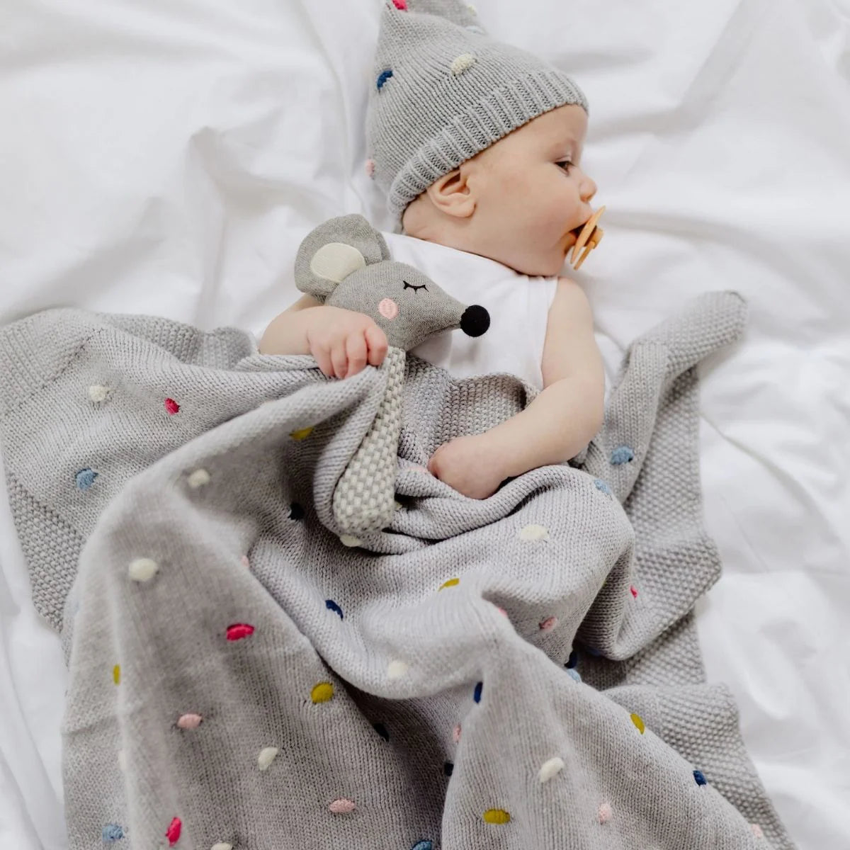 Baby wrapped in a gray blanket with colorful polka dots, wearing a matching hat and holding a toy, on a white background.