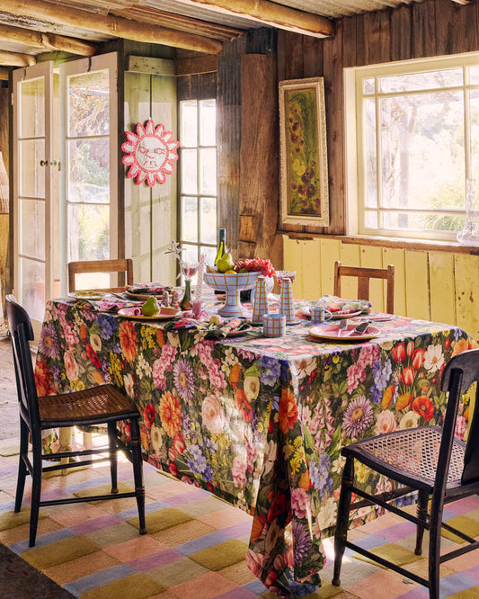 A rustic dining room scene with a floral tablecloth draped over a rectangular table set with plates, glasses, and chairs. The room has wood-paneled walls, large windows, and a colorful checkered rug.