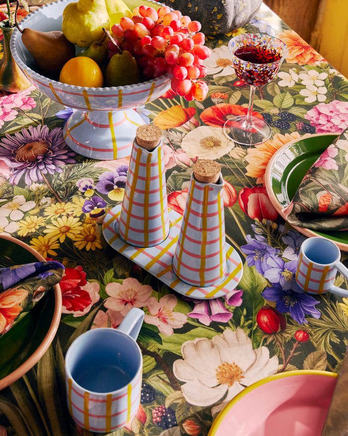 Colorful table setting with floral tablecloth, teapots, cups, and fruit on a patterned surface.