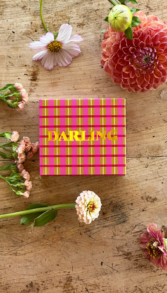 Boxed soap bar sits on timber table with flowers sitting around box. Box is pink and mustard plaid design. Gold leaf Darling written on box.