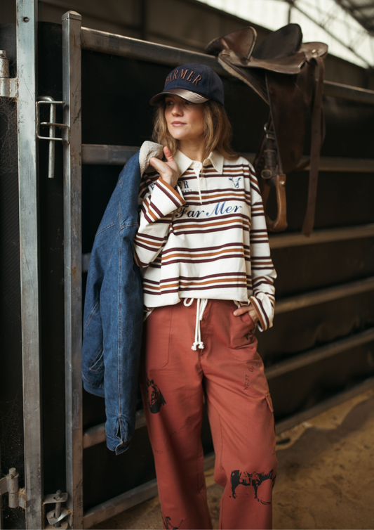 A woman stands in a rustic barn or stable doorway wearing a striped long-sleeve polo shirt, rust-colored cargo pants with horse graphics, a blue denim jacket, and a navy baseball cap with 'Farmer' embroidered on it.