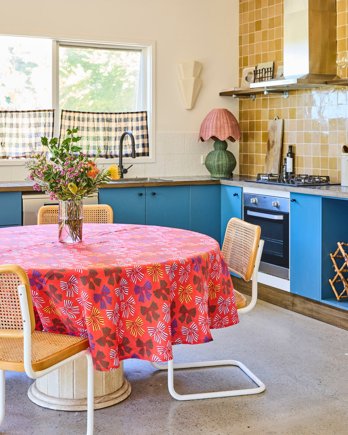 A round dining table covered with a bright coral-red floral tablecloth, surrounded by woven-back chairs in a real kitchen setting with blue cabinets, a tiled wall, and a window.