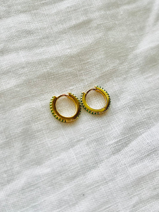 Gold hoop earrings with green crystal around front of each hoop, laid on a light fabric background