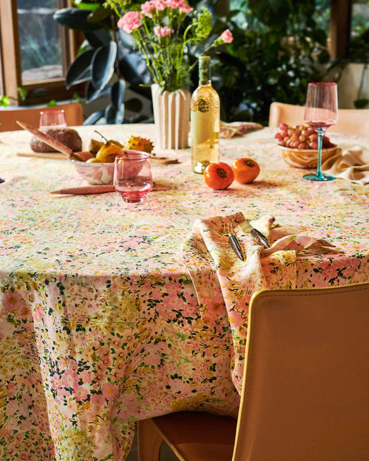 Pink, green and yellow floral linen tablecloth laid out on round table, with scattered wine glasses, fruit and vase with flowers. N