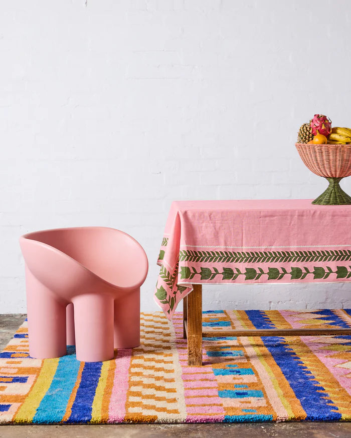 A pink sculptural chair sits on a colorful striped rug next to a pink table with a patterned tablecloth and a fruit bowl, set against a white brick wall.