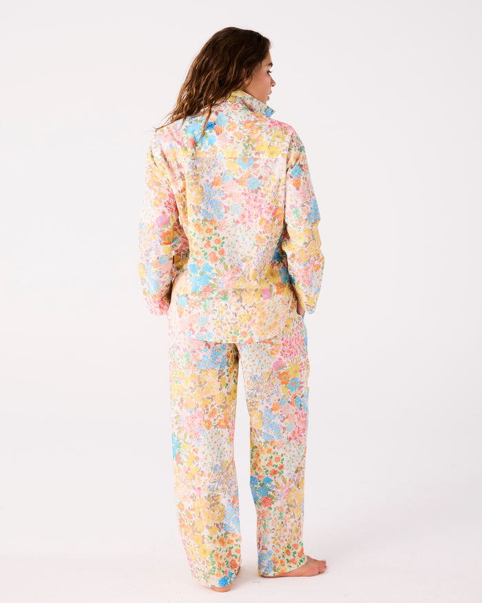 A woman stands barefoot against a plain white studio background, viewed from the back, wearing a matching long-sleeve fleece jacket and pants set in a pastel floral print.