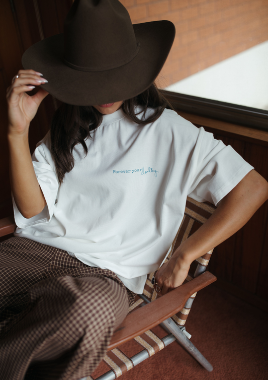 Person wearing a white t-shirt with text and a dark hat, sitting on a wooden chair.