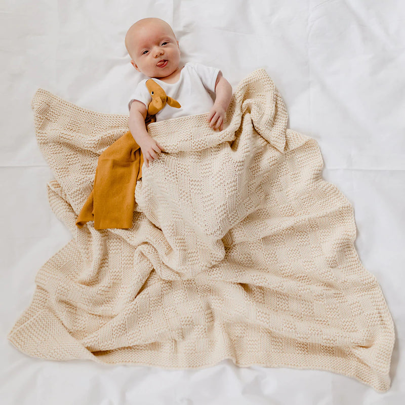 Baby lying on a white blanket with a beige knitted blanket and toy deer.