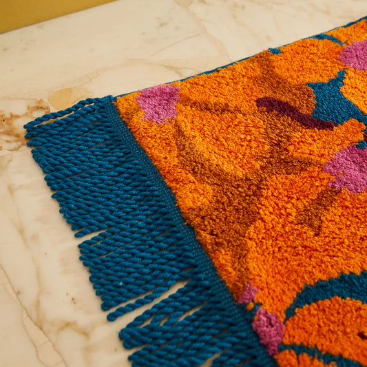 Close-up of a colorful woven bath mat with blue and orange patterns, blue fringing detail, on a light wooden floor.