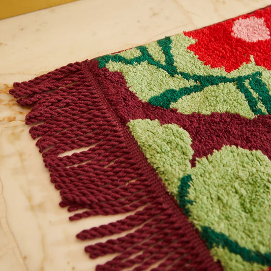 Close-up of a textured bath mat with red and green patterns, red fringing detail along side of bath mat, on a wooden surface