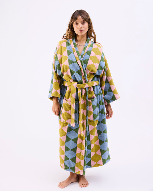 A woman modeling a long bathrobe with a colorful geometric diamond pattern in olive green, dusty blue, and pink, set against a plain white studio background.