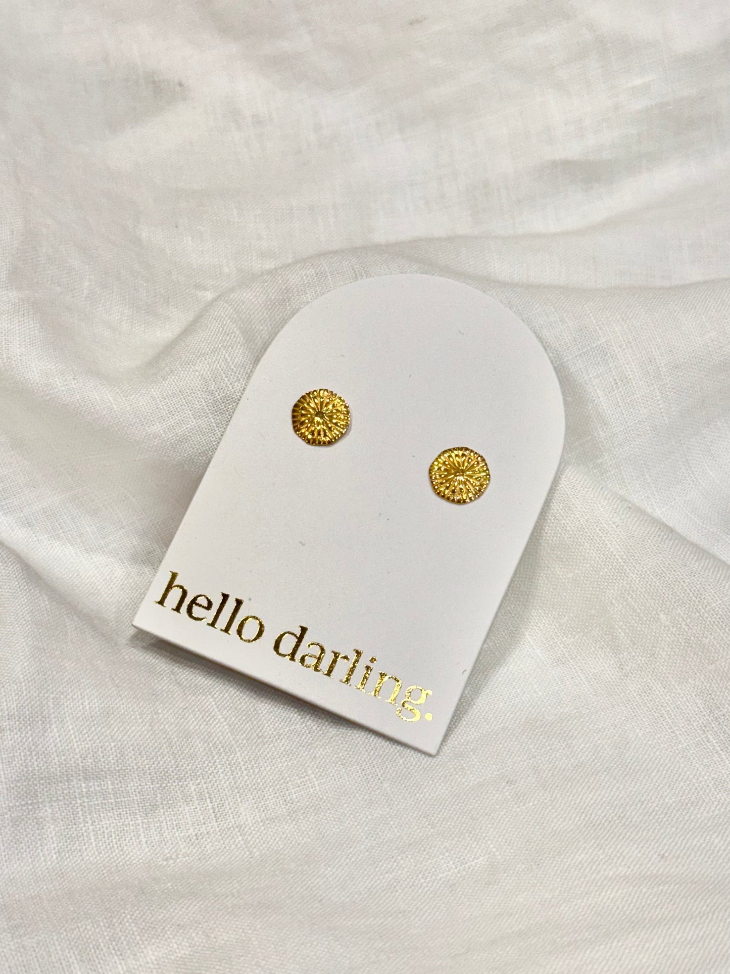 Gold earrings on a 'hello darling' branded card against a white fabric background
