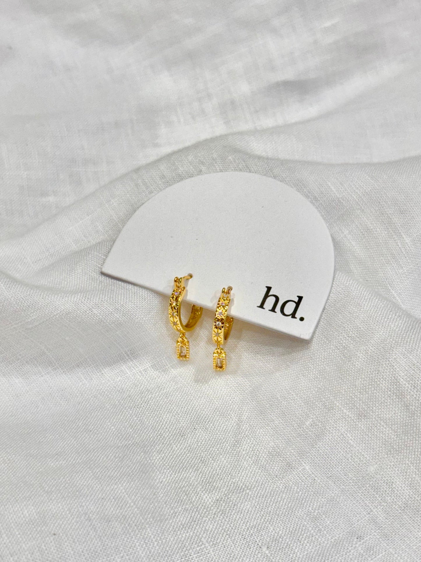 Gold earrings on a white card with 'hd.' text, placed on a white fabric background