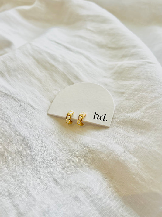 Gold earrings with three small pearls on a white card with 'hd.' text, placed on a white fabric background