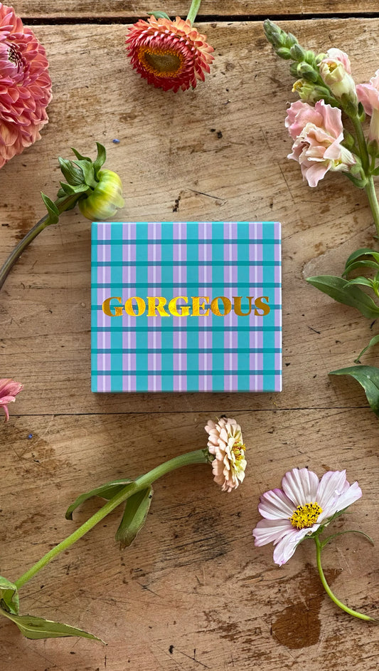 Boxed soap bar with gorgeous written across front in gold leaf, box packaging is plaid pattern in blue and lilac. Timber background with flowers sitting around box.