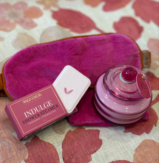 Pink eye mask, shower steamer, and pink candle jar on a floral fabric background