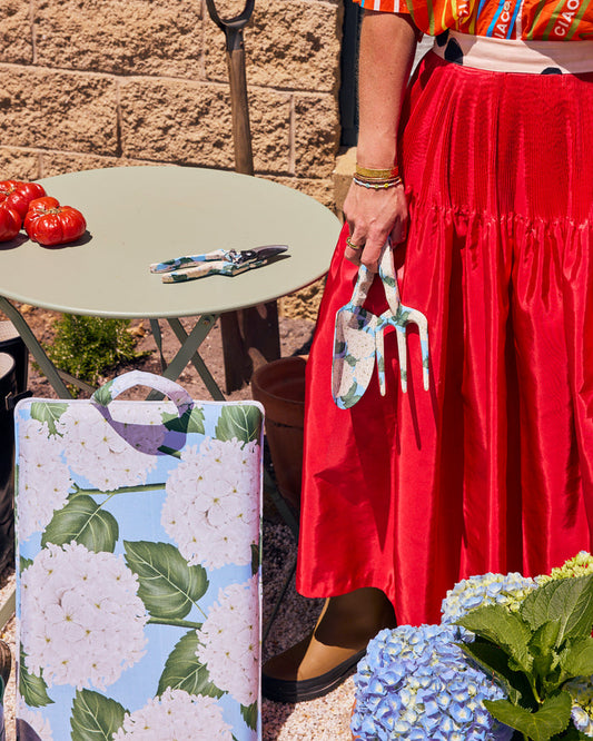 A person in a bright red skirt holds a small gardening trowel and cultivator next to a decorative metal garden tote with a white floral print on a light blue background. The scene is set outdoors in front of a stone wall with a small table holding tomatoes and pruning shears.