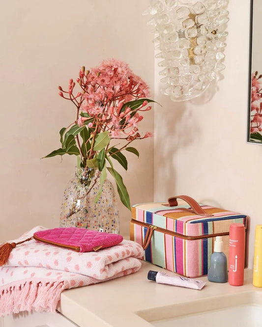 Stripe toiletry case sits on vanity surrounded by pink polka dot towel, various skincare products, glass vase with pink australian native flowers.
