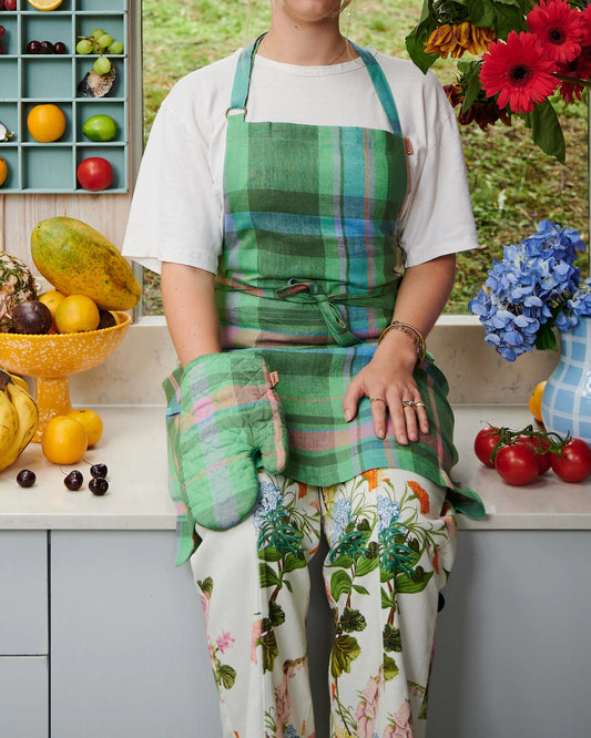 Woman sits on kitchen bench surrounded by fruit and jug filled with flowers, she wears apron and matching oven mitt in green base, with blue pink and lilac tartan pattern oven mitt.