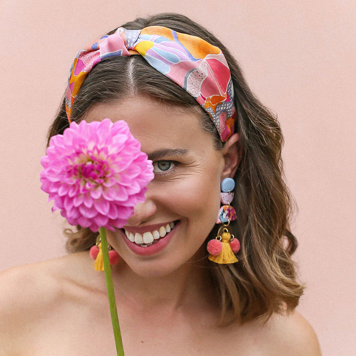 Woman wears silk floral headband, hand drawn design. Wears matching colourful earrings and hold pink flower in front of face.