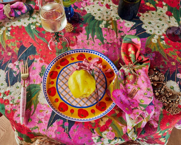 Bush blossom tablecloth laid out on table, styled with tomato pattern plates, matching napkins, wine glass and cutlery. Colours featured within tablecloth include pink, red, green and cream florals.