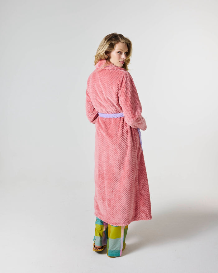 A woman wearing a pink textured bathrobe with a matching waist tie, shown from the back against a plain light gray studio background.