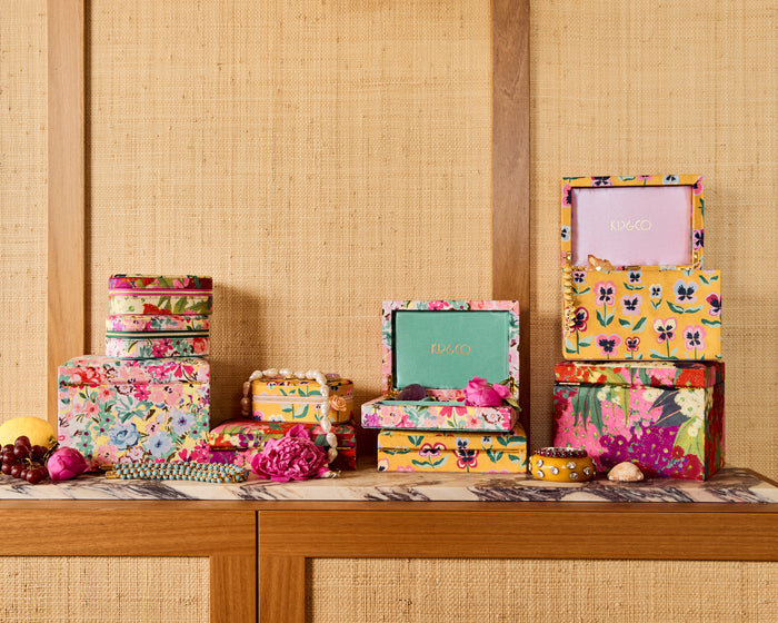 A collection of colorful floral and patterned storage boxes and small decorative containers arranged on a wooden surface against a woven beige wall. The boxes feature vibrant multicolor prints including pink, yellow, green, and blue, with one box showing a light pink interior and another a mint green interior.