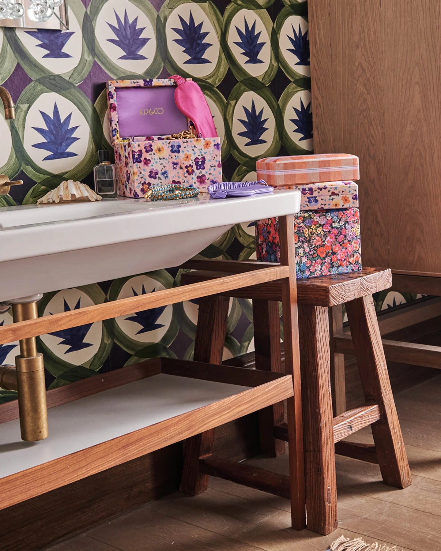 Bathroom with exposed timber shelving and timber stool. velvet jewellery boxes sit on stool and another on vanity with jewellery cascading out of jewellery box. Patterned green and blue wallpaper.
