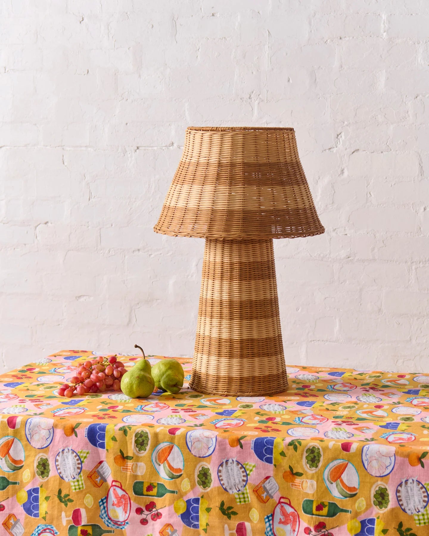 Supper pattern tablecloth laid out on table, with wicker lamp sat on top of table and two pears and group of grapes sat next to lamp. White wash brick wall in background.