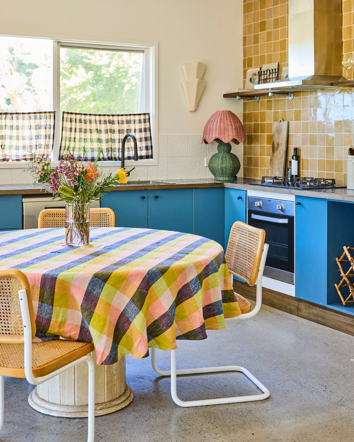 A round dining table covered with a pastel plaid tablecloth, surrounded by woven-back chairs in a bright kitchen with blue cabinets and yellow tile.
