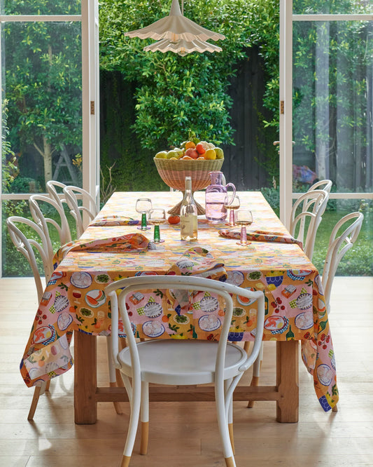 Supper tablecloth laid out on timber table with white chairs sitting around table and big open white doors in background.
