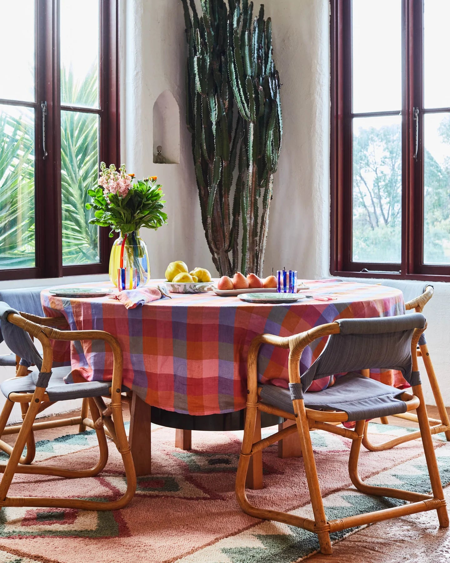 Round table with pink, purple, blue and red tartan tablecloth laid out. Sat on top table is glass vase and flowers, fruit on platters.