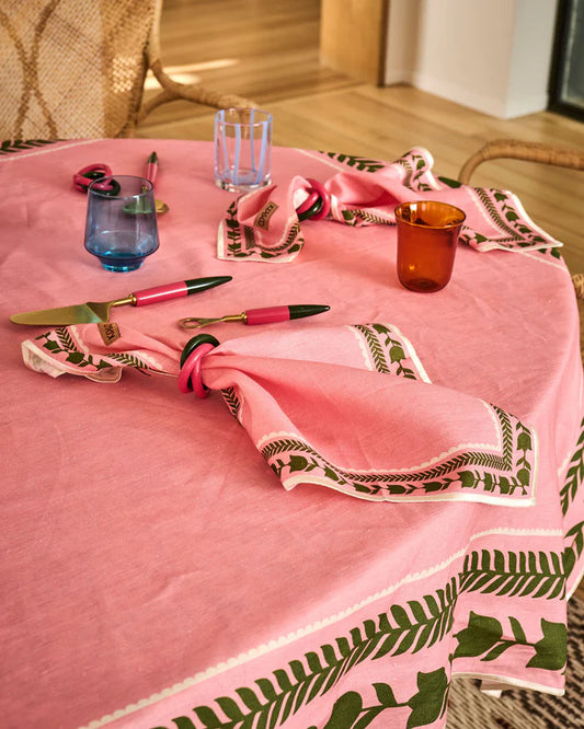 A pink tablecloth with a green botanical leaf border pattern, styled on a wooden table with matching pink napkins, cutlery, and glassware.