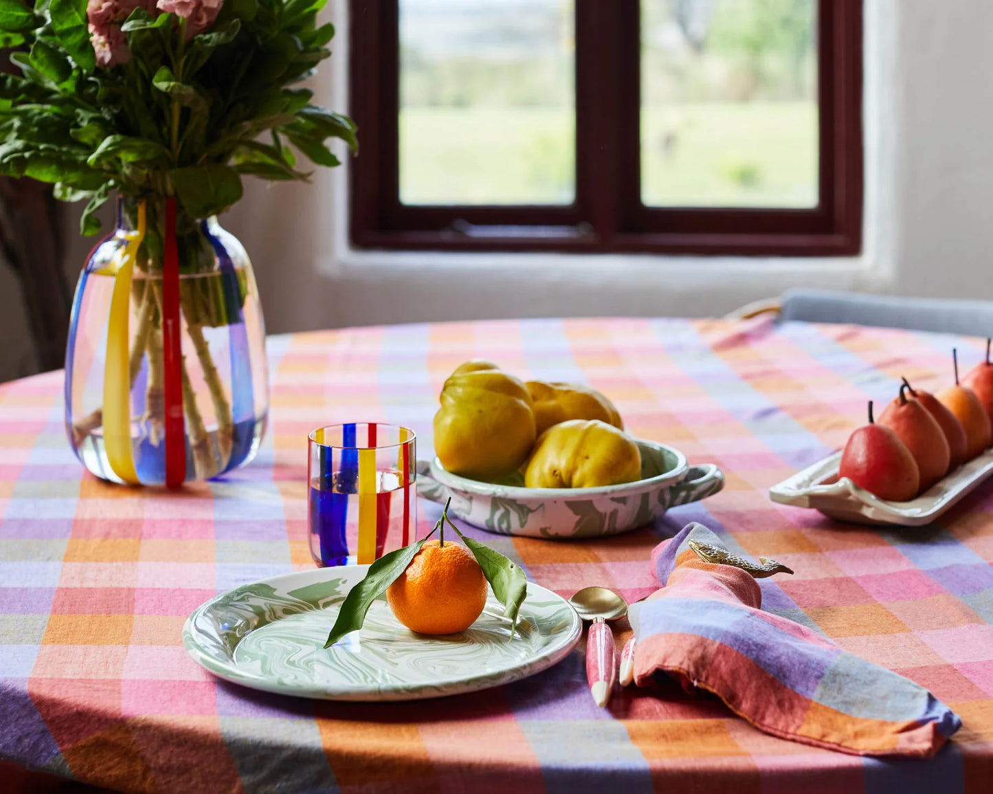 Styled table with stripe vase and flowers, matching water glass and plates with various fruits. Laid out is rainbow tartan tablecloth and matching napkin with fish napkin holder.